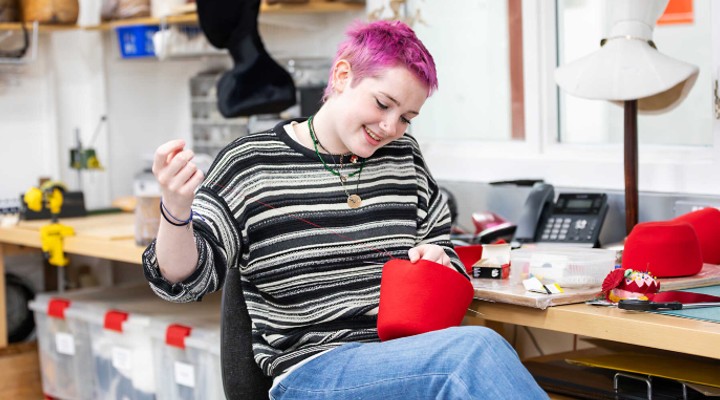 A costume apprentice works on a red, fez-like hat
